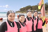 Five young women dressed in pink and black wetsuits smile as they stand in a row on a sunny beach.