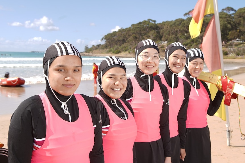 Five young women dressed in pink and black wetsuits smile as they stand in a row on a sunny beach.
