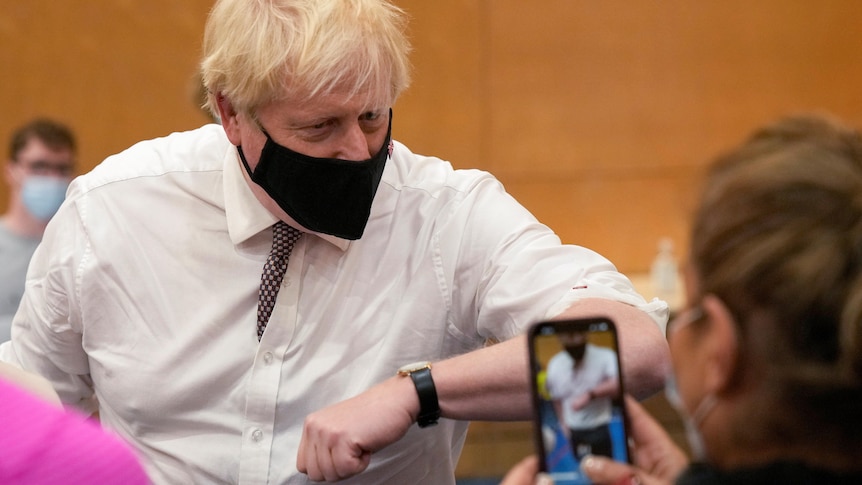 Boris Johnson wearing a black mask and a white shirt and tie elbow bumps a person at a vaccination centre as someone films