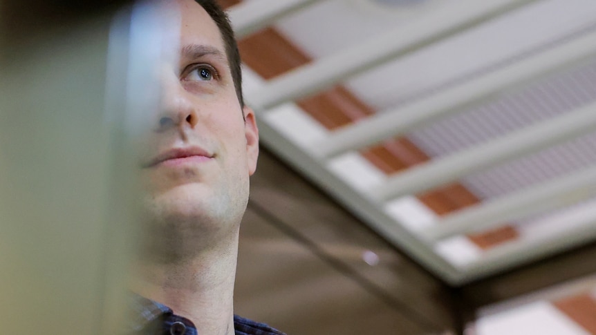 Evan Gershkovich, wearing a plaid shirt, looks up, half his face covered in glass reflection as he stands inside a glass box