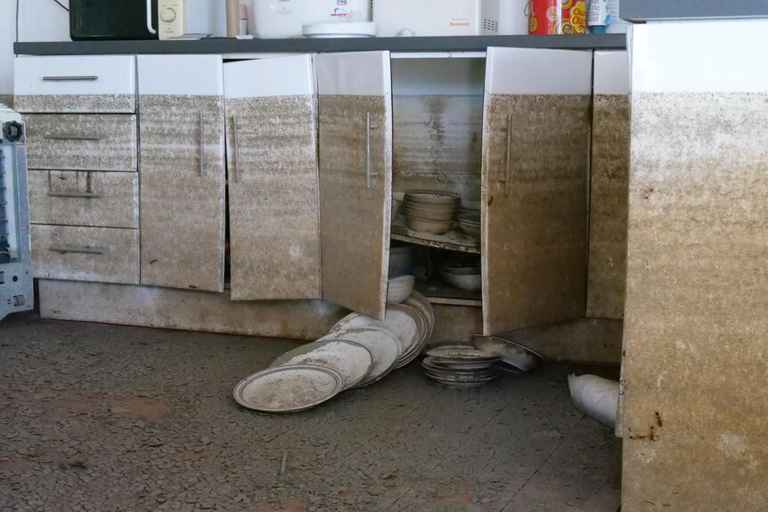 Mud covered plates spill out of cupboards in a kitchen with a water mark almost up to the benchtops. 