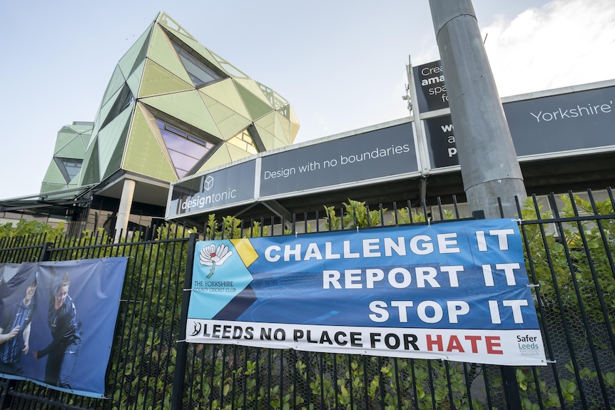 A banner hanging on a fence outside a stadium reads "Challenge it. Report it. Stop it"