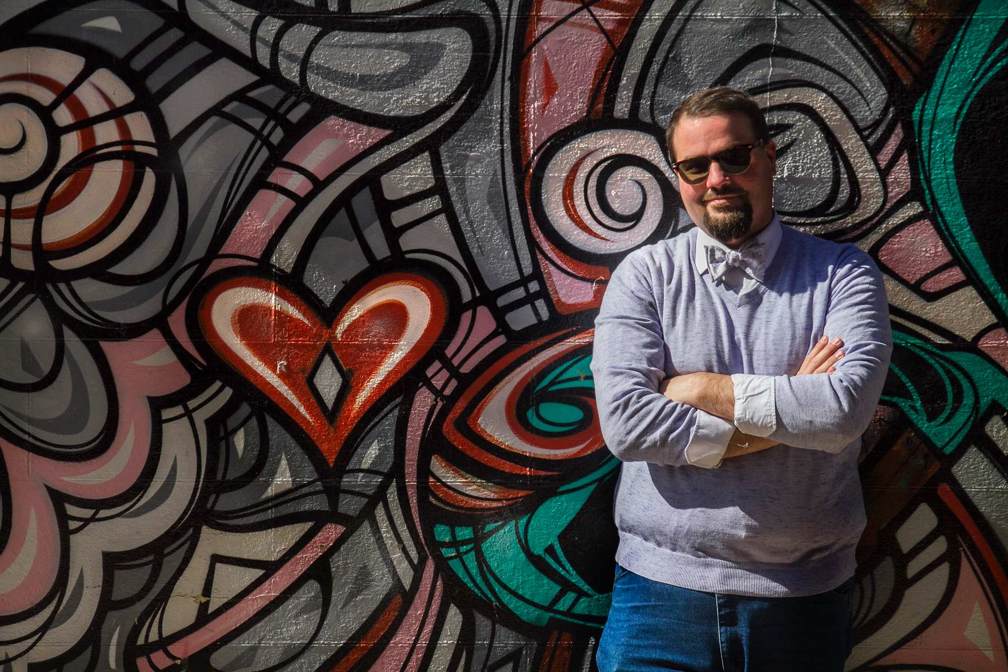 A man in sunglasses and a bow tie leans agaisnt a mural painted wall