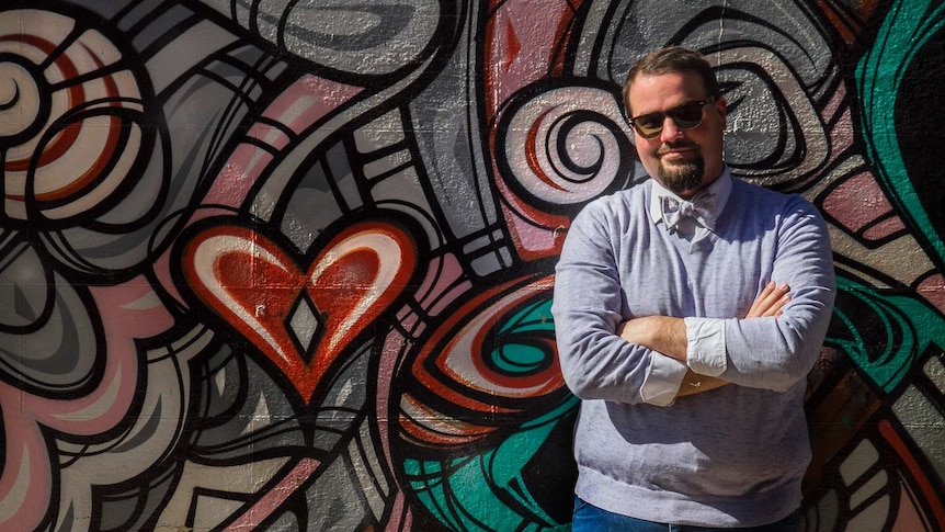 A man in sunglasses and a bowtie leans agaisnt a mural painted wall.