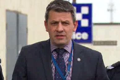 A dark-haired man in a dark suit speaks to the media on a grey day.