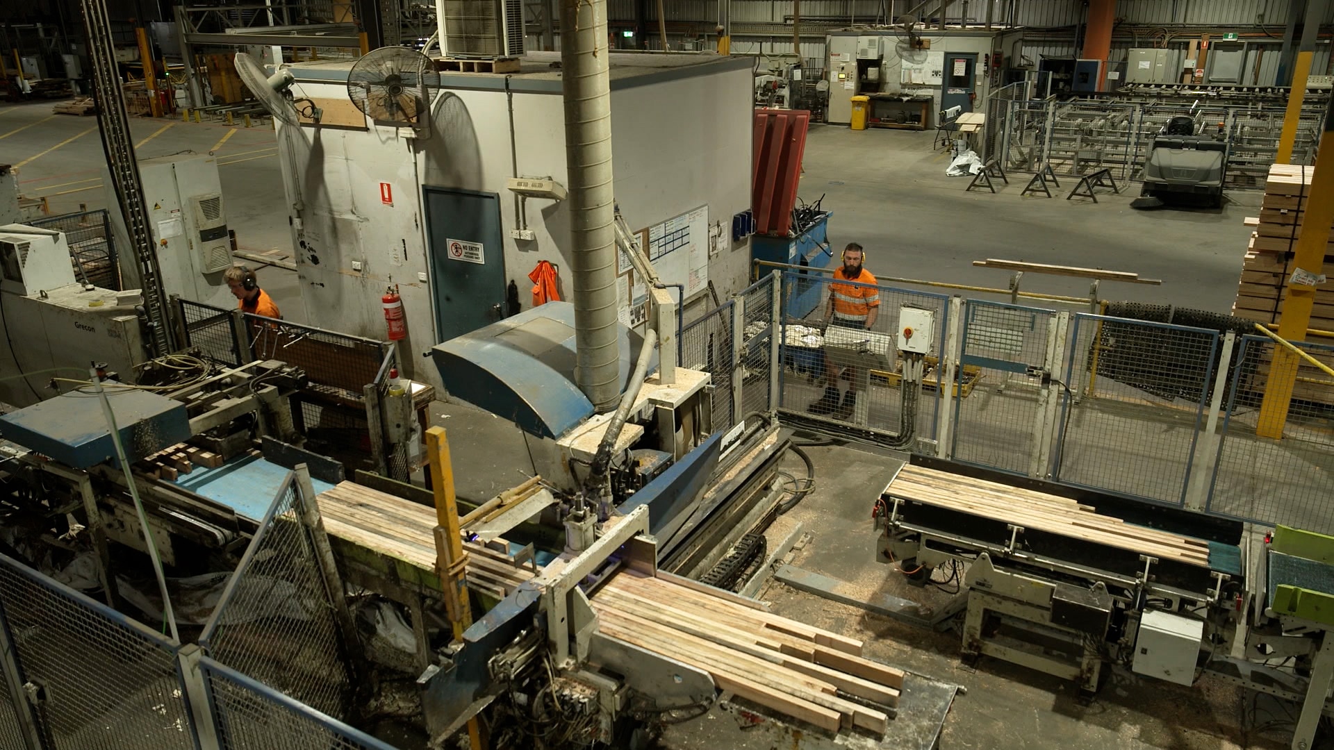Lengths of timber sit on conveyor belts of large machines in a factory. A couple of workers can be seen standing nearby.