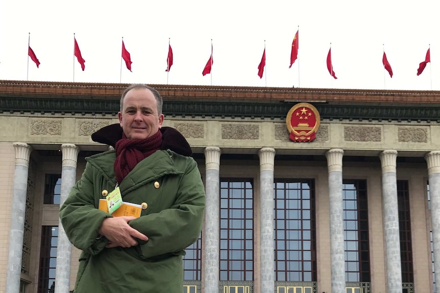 A man in a green jacket stands outside a building with columns and red flags on the top and a red and gold emblem