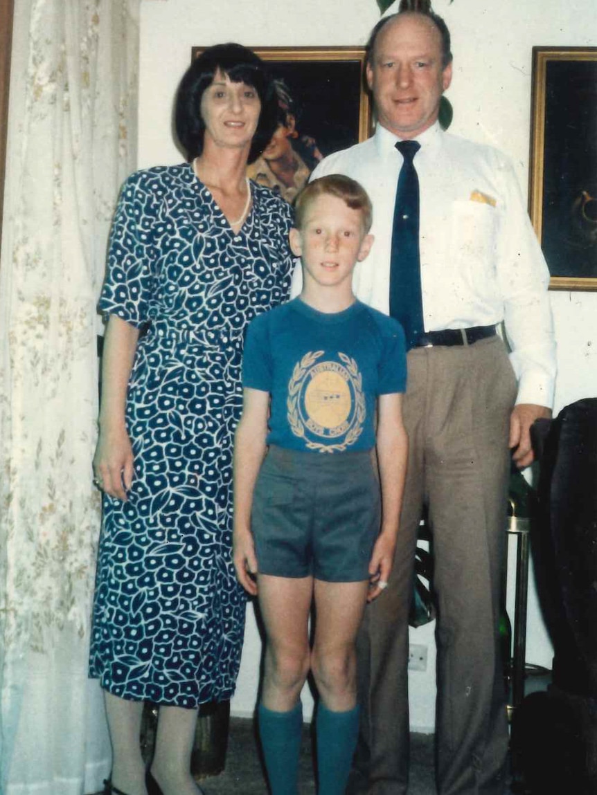A boy stands in front of a man and woman, all look at camera.