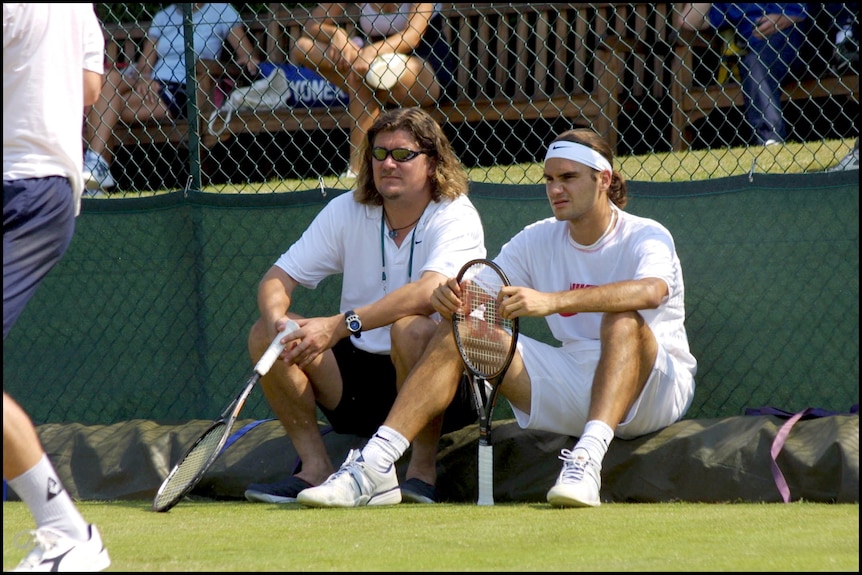 Two men sit on the side of a tennis court.