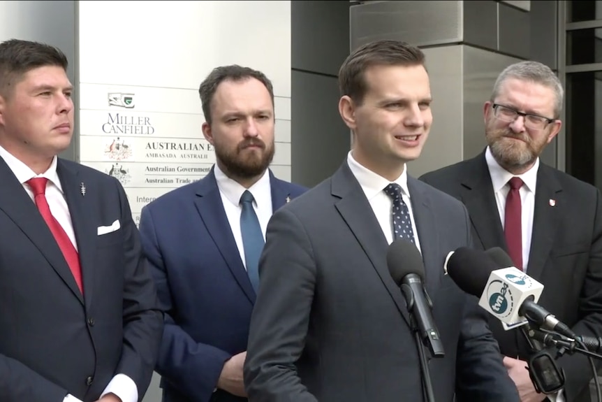 Four men in suits speaking at a press conference