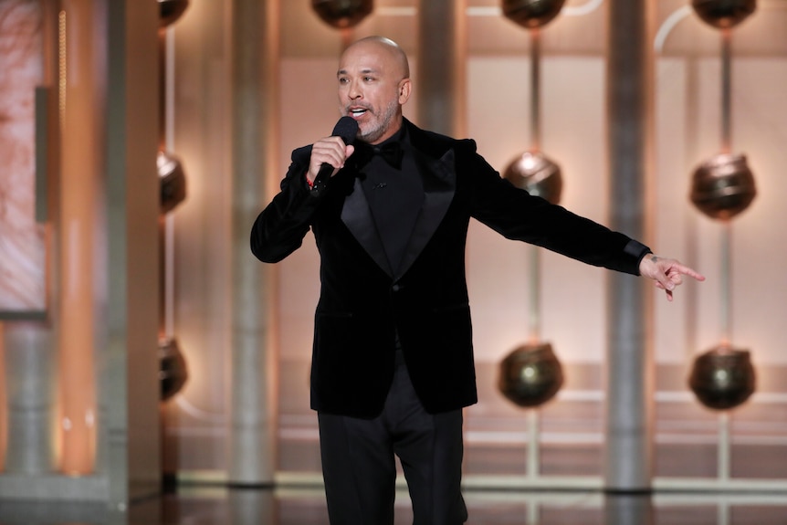 Bald man in a black suit holding up a microphone on stage