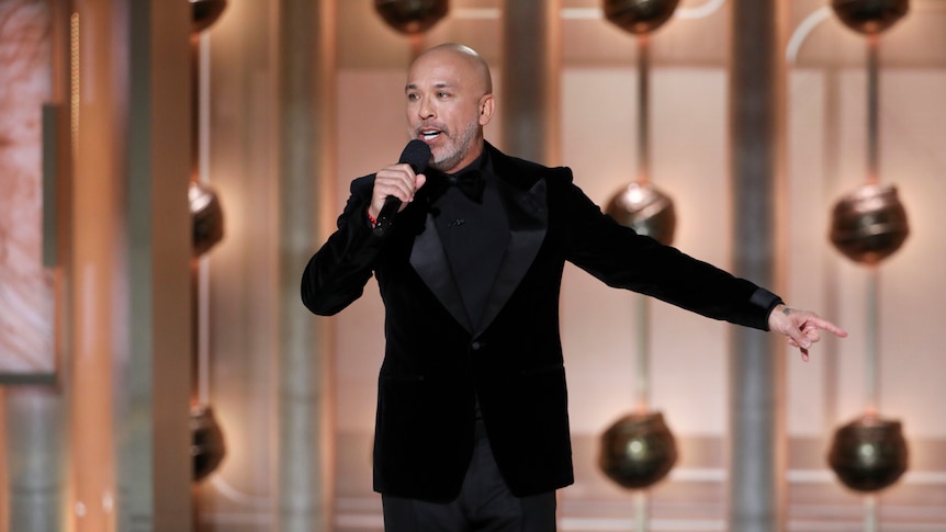 Bald man in a black suit holding up a microphone on stage