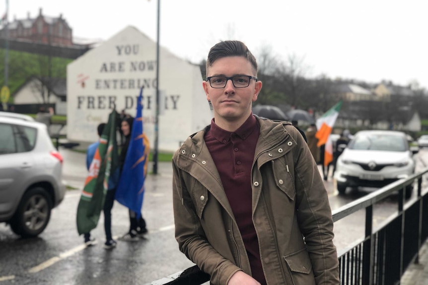 Patrick Gallagher wears a brown coat and maroon polo shirt as he stares at camera with slight smile and black glasses by road.