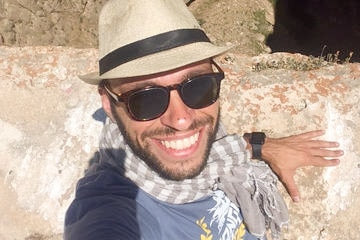A man wearing a hat and sunglasses smiles as he sits on a rock.