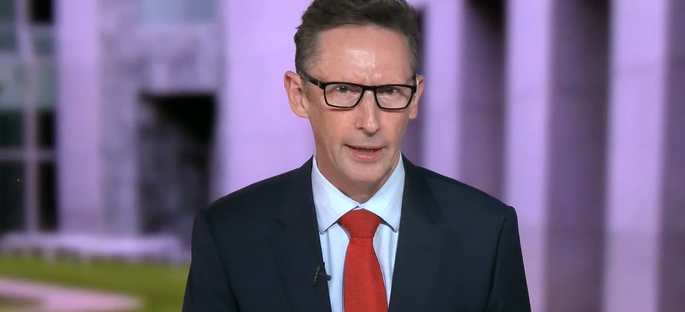 A middle-aged man in a suit and tie and glasses gives a TV interview in front of a backdrop of Canberra parliament house