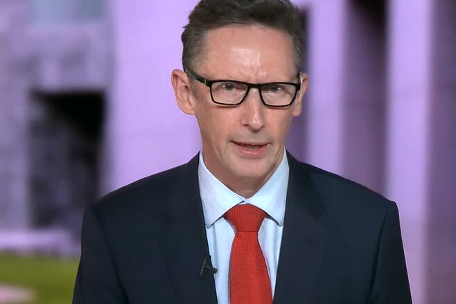 A middle-aged man in a suit and tie and glasses gives a TV interview in front of a backdrop of Canberra parliament house