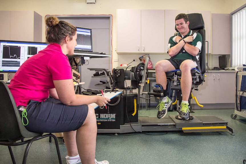 A woman with a clipboard and computer monitors in the background and a high school boy sitting strapped into a gym type chair