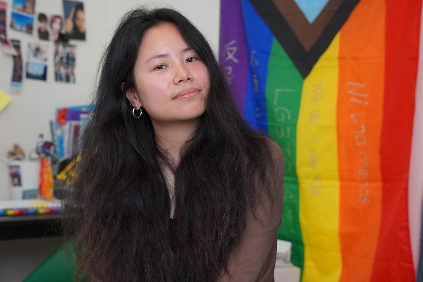 A Chinese person sits in front of a rainbow flag. 