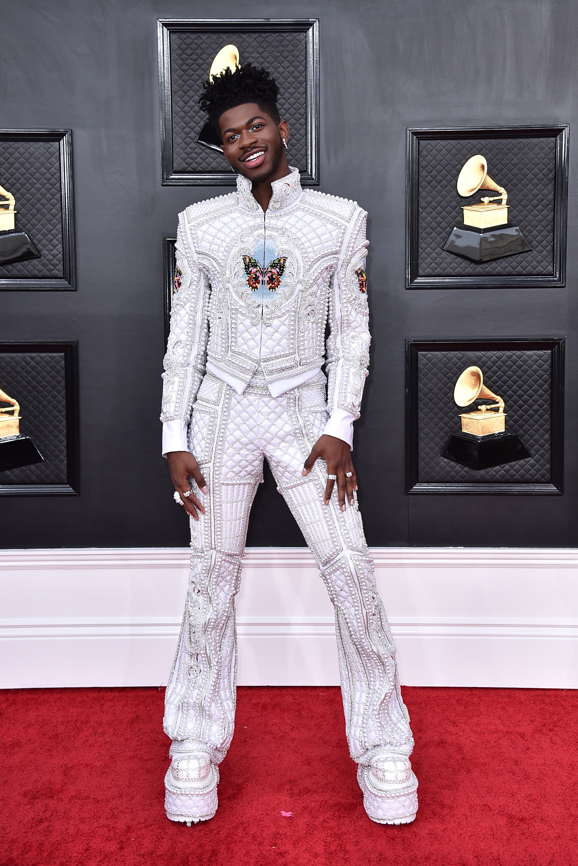 lil nas x wearing a white heavily embellished suit with a butterfly detail and platform matching white boots on the red carpet