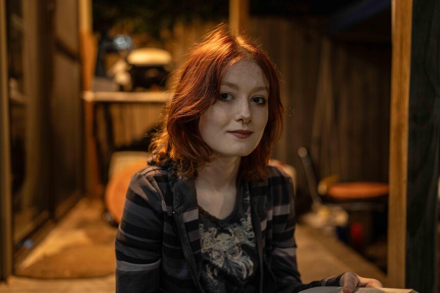 A teenage girl sits outside at night, looking at the camera with a slight smile. A light illuminates the house behind her.