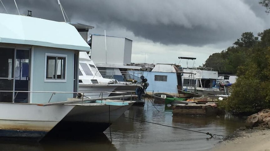 A group of ageing boats, including houseboats, anchored close to a small beach under threatening storm clouds