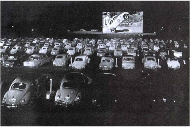 Rows of old cars line up in front of a large outdoor screen with an image of a car-related movie on it.