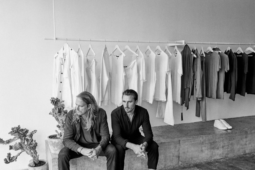A black and white image of two men in shirts and pants, with clothes hanging on a rack behind them in an open room.