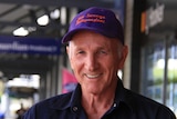 A man with a purple and red cap smiles at the camera as he stands on a footpath lined with shops.