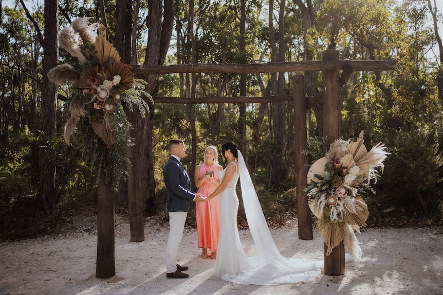 A bride and groom say vows with a celebrant in front of a scenic bush backdrop.