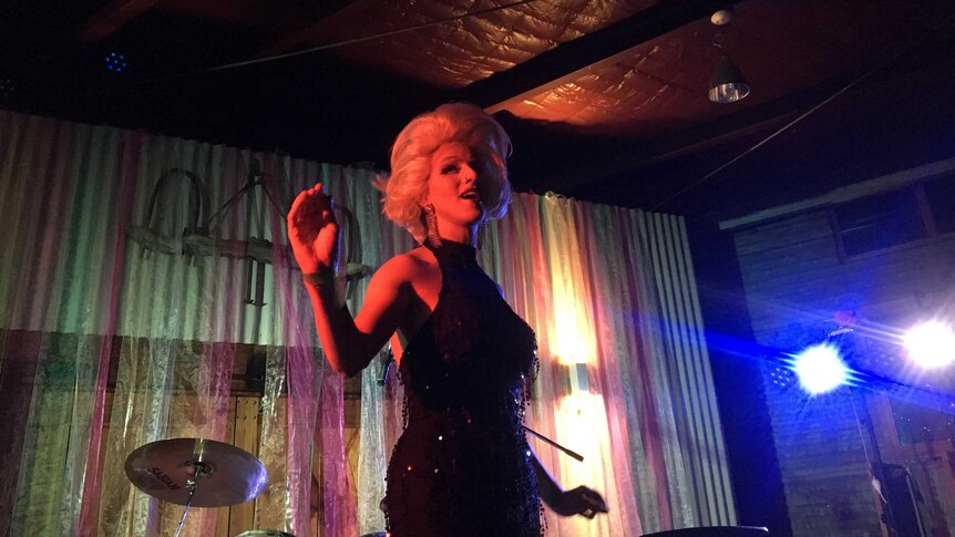Alicia Zaman performs on stage, wearing a black sequined gown, blond wig, with red light shining on her face