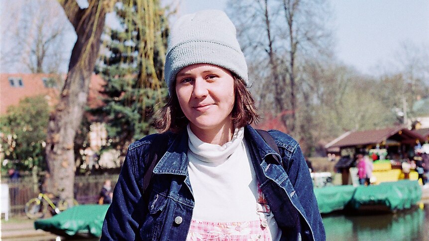 A woman stands in a park wearing a denim jacket and grey beanie.