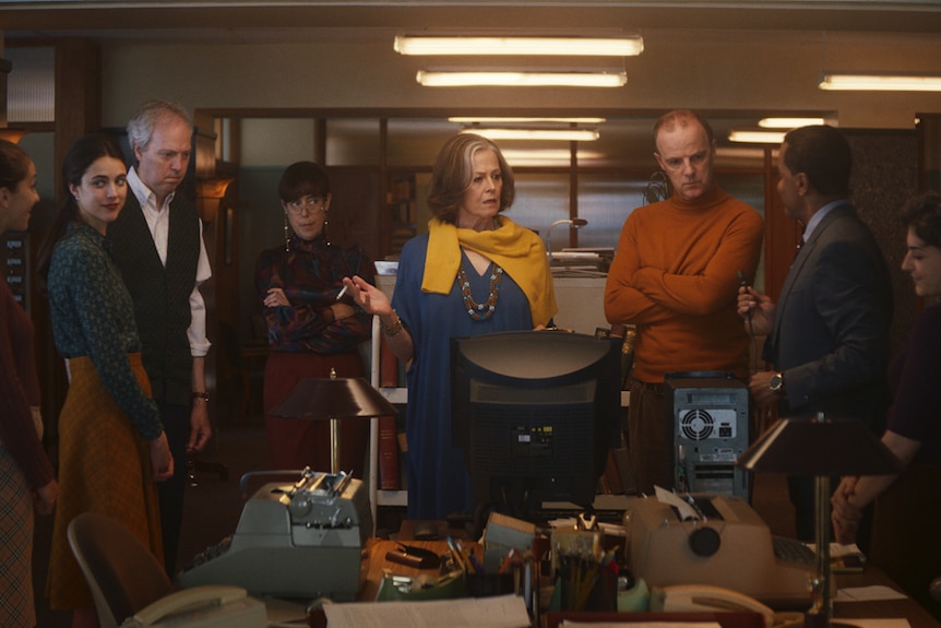 Dimly lit office with group of employees standing around desk with 90s computer on it, Sigourney Weaver standing in front of it.