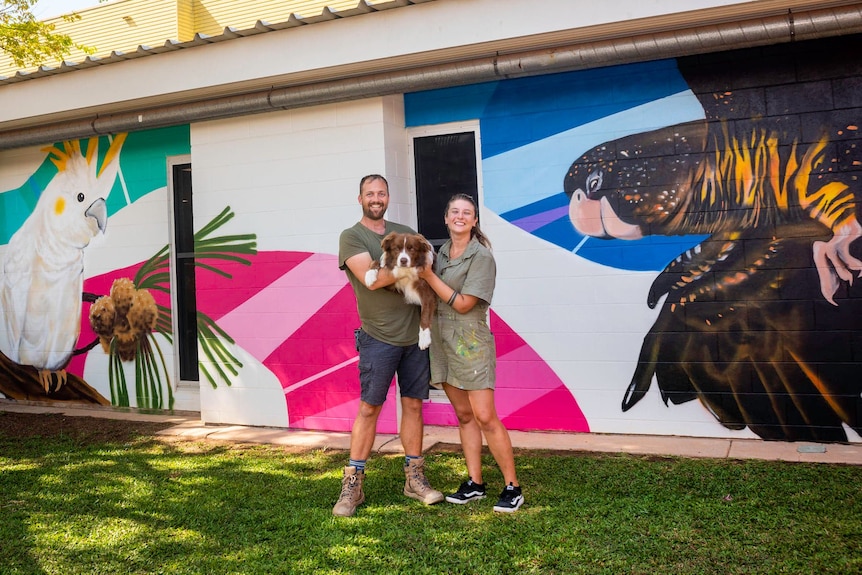 a man and a woman hold a dog in front of a colourful mural