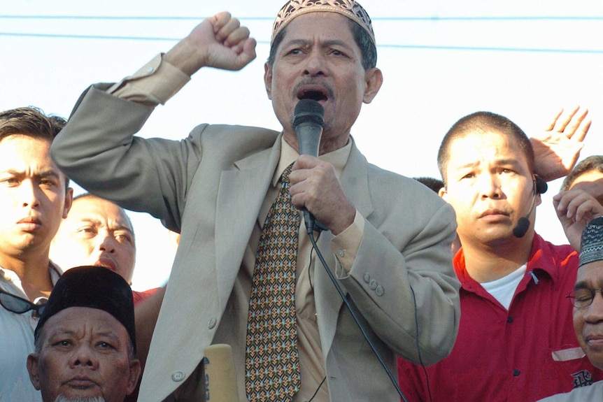 Misuari wearing a brown suit speaks passionately into a microphone, one fist held aloft, at a rally.