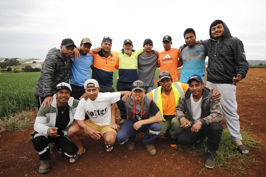 A group of 13 men stand arm in arm as they pose for a group photo in a farm field.