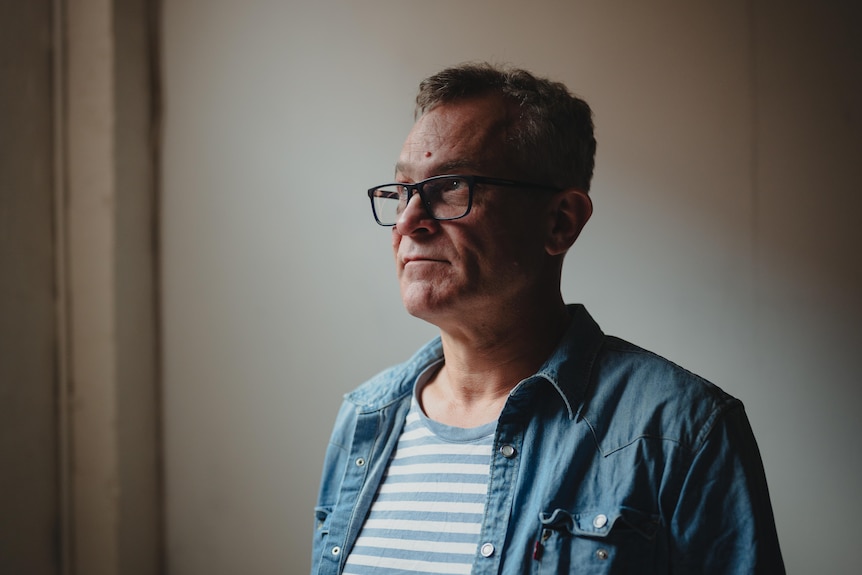 A man wearing glasses stands in a room, looking up slightly towards a source of light. Part of his face & the room is in shadow.