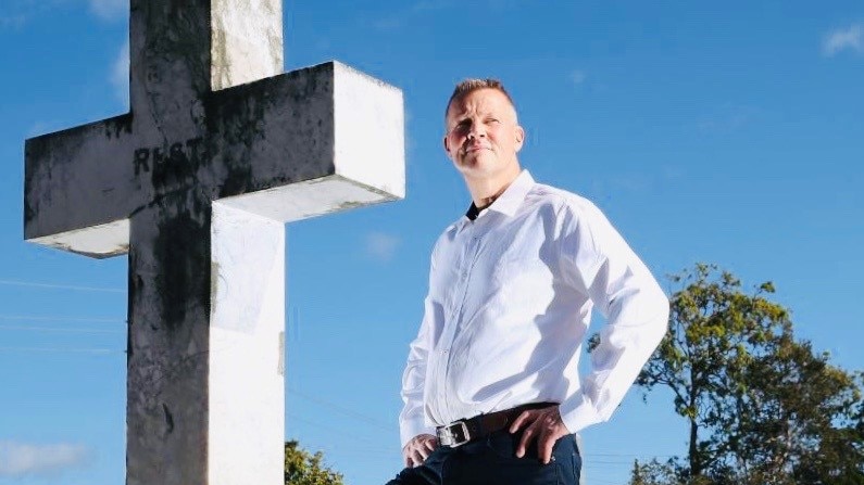 Man in white shirt and black pants stands looking serious beside a cross in a cemetery.