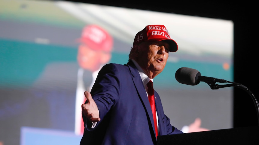 A man in a suit and a red baseball cap gestures with wide arms as he speaks to a crowd with a large video wall behind.