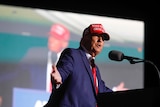 A man in a suit and a red baseball cap gestures with wide arms as he speaks to a crowd with a large video wall behind.