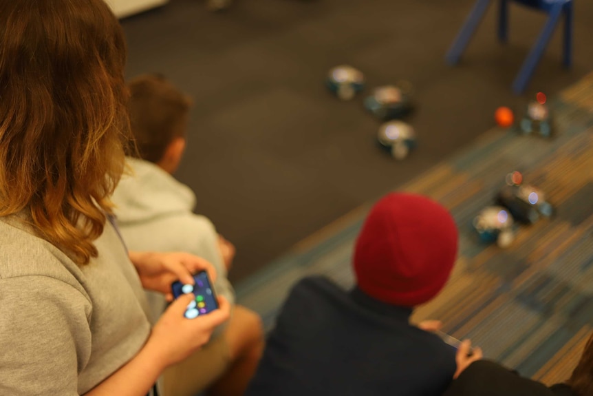 Girls hold iPhone to control robots on carpet in class.