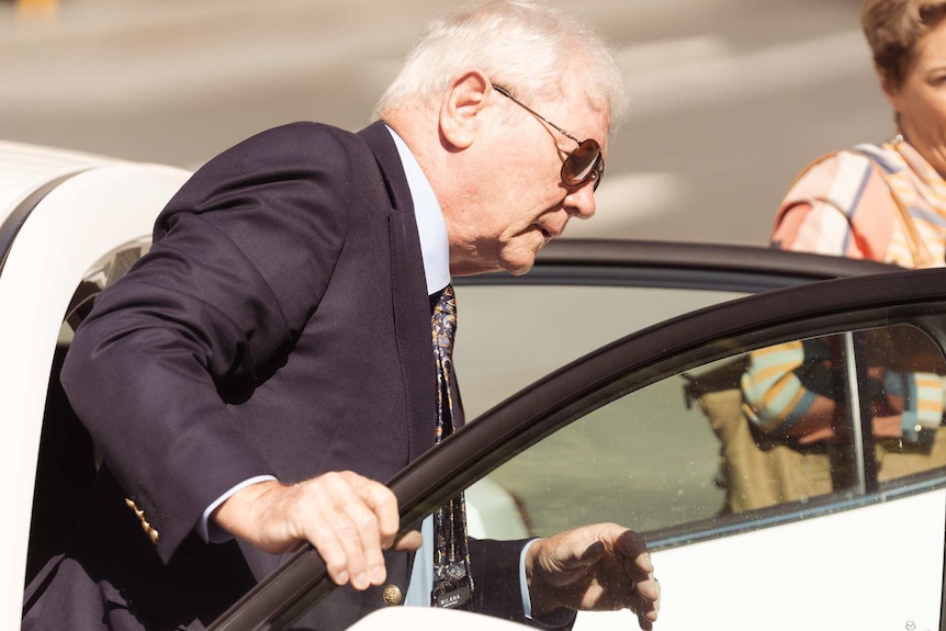 Denis Glennon steps out of a white car wearing a suit and sunglasses, with a woman behind him.