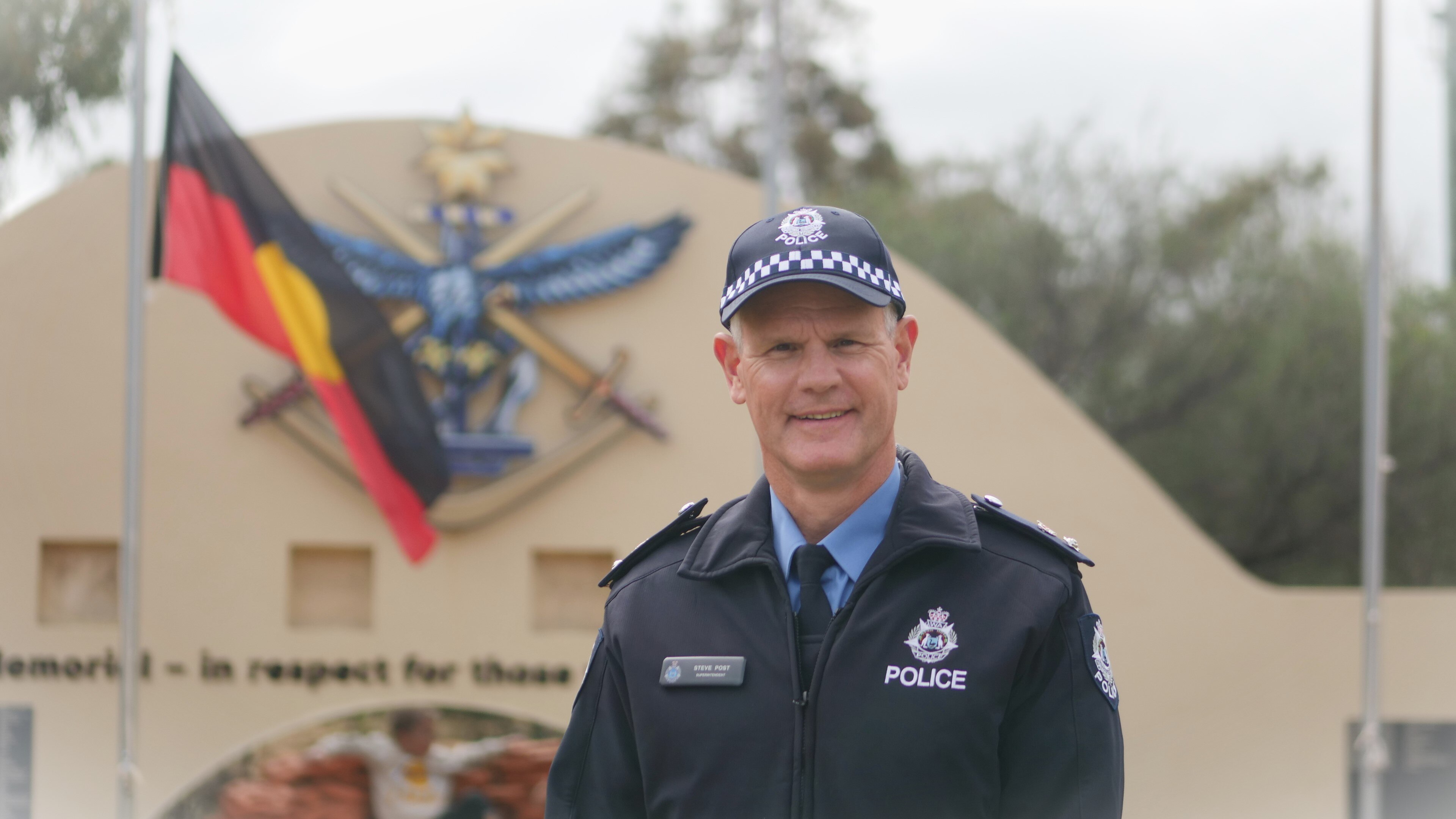 A man in a blue police uniform stands off right smiling with an Aboriginal red, black and yellow flag in background. 
