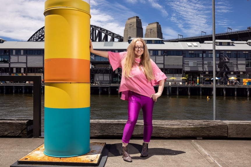 A blonde white woman in her 40s wears a vibrant pink outfit and stands on a dock, leaning against a colourful pylon.