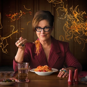 A woman seated at a table in front of a spaghetti and sauce swirled wall, holding a fork with spaghetti above a plate of pasta