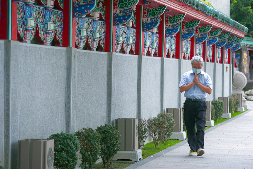 A man in a face mask holds his hands together in prayer near a temple wall