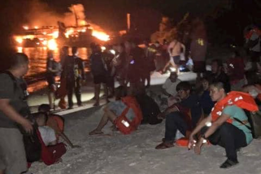 People in life vests sit on shore with burning ship in background