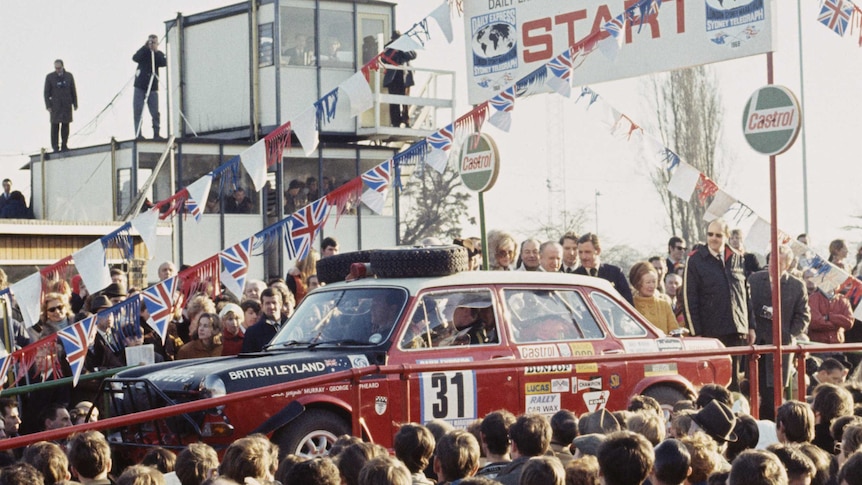 A red rally car sits at the start of a motor race surrounded by people.