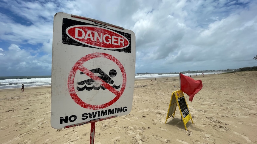 A 'Danger no swimming' sign on a Sunshine Coast beach, with blue but cloudy sky.