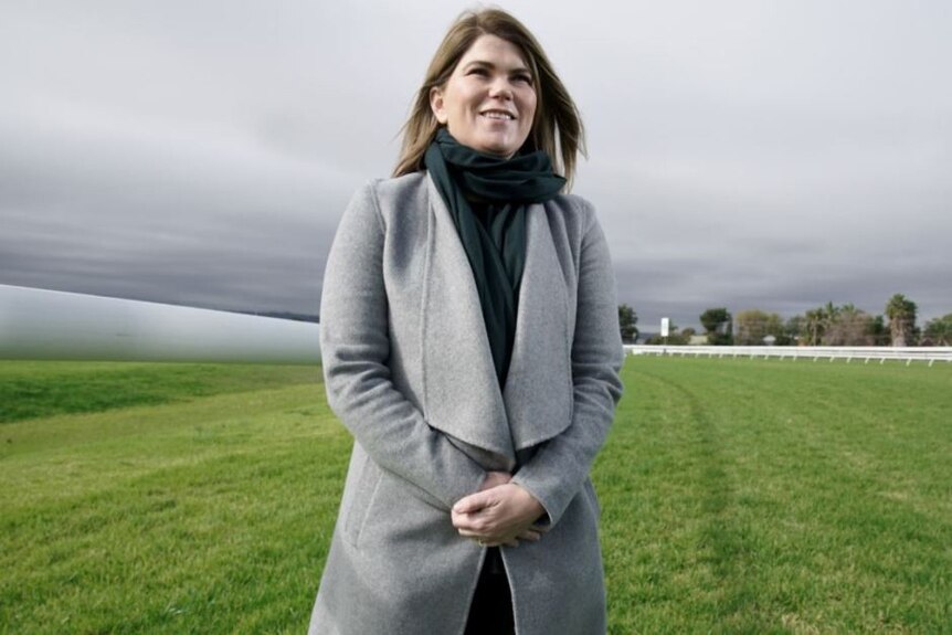 A woman in a grey coat and black scarf stands on green grass on a racing track.