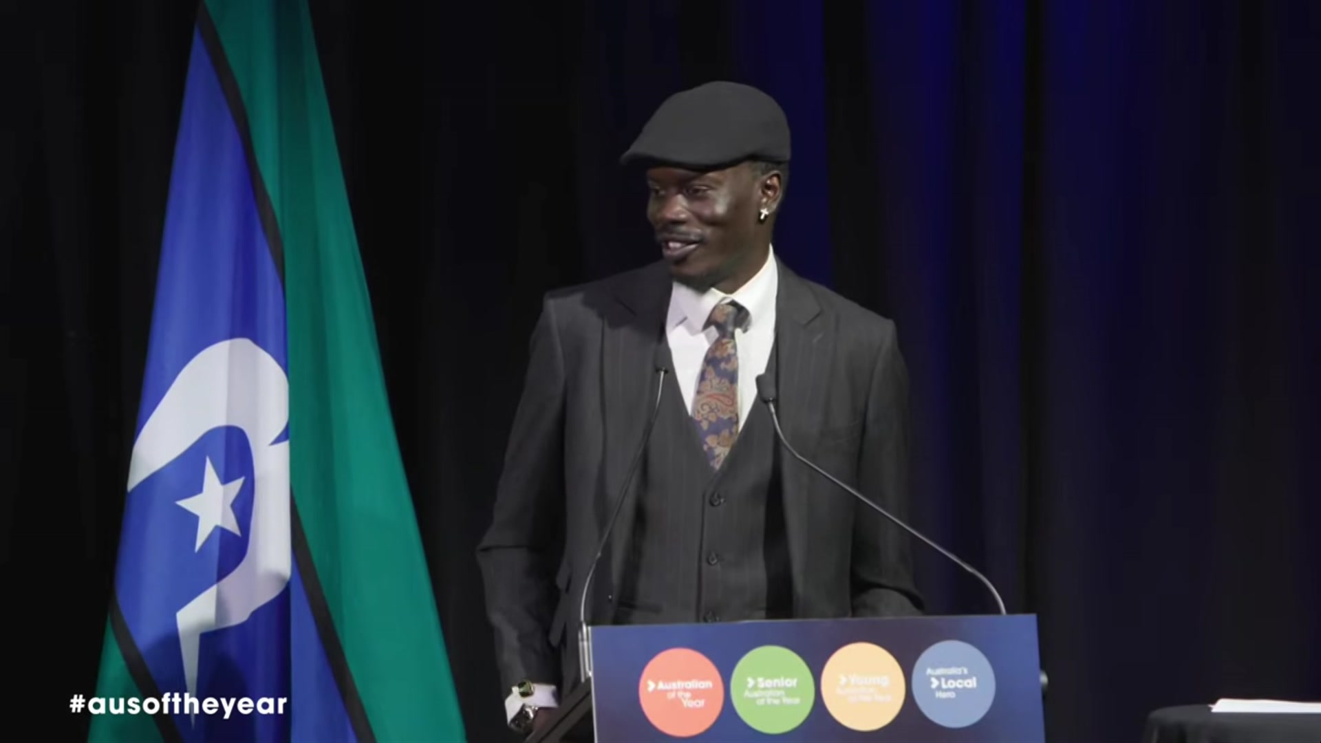 A man in a dark suit jacket, vest, cap, white collared shirt and tie stands on a stage smiling.